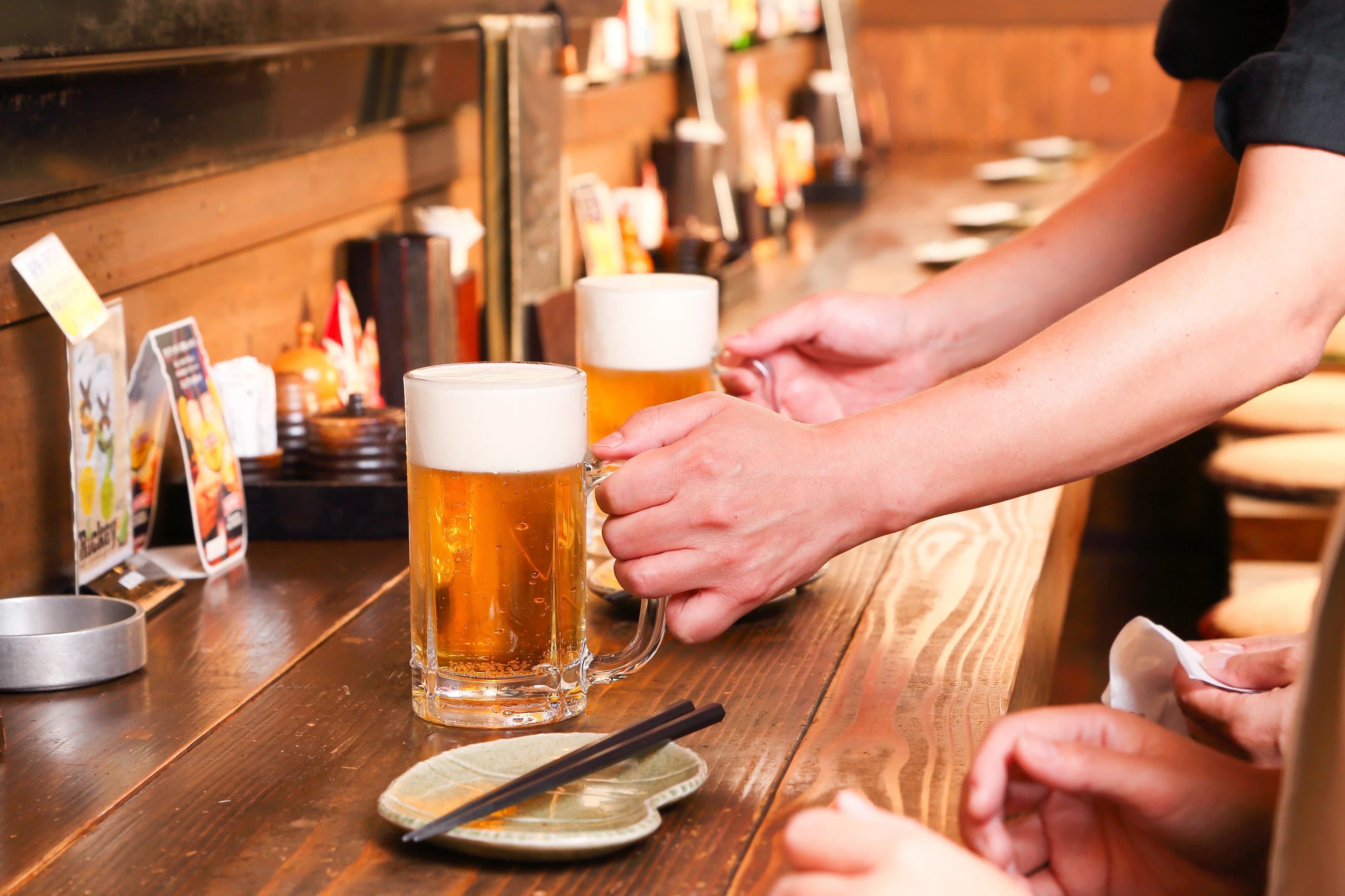 居酒屋のカウンターで、店員が客に生ビールのジョッキを手渡しているところ。木製カウンターには泡の立った生ビールがもう一杯と、箸をのせた小皿が置かれている。
