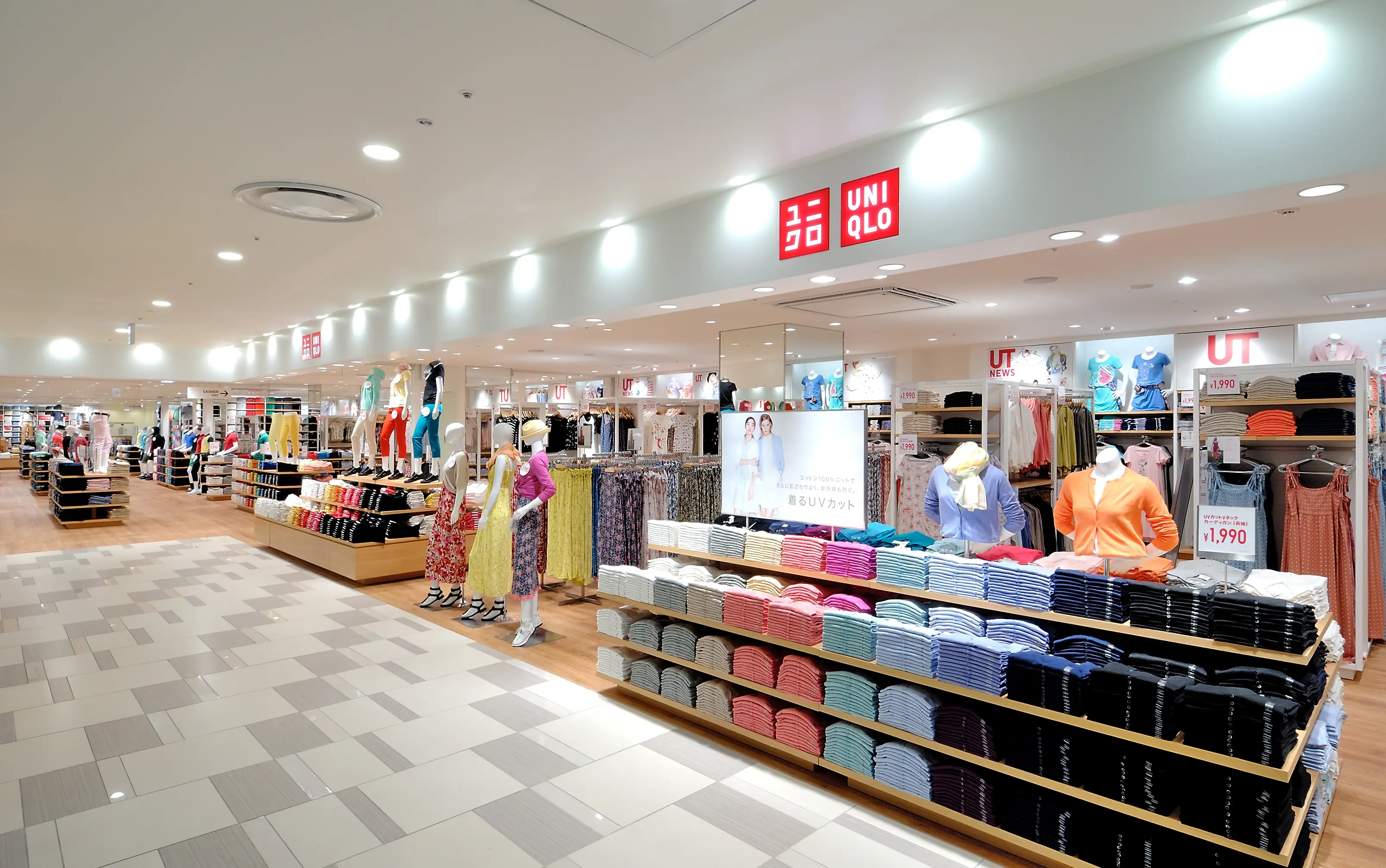Storefront of a UNIQLO shop inside a shopping mall, with shelves of color-sorted folded T-shirts, polo shirts, and mannequins.