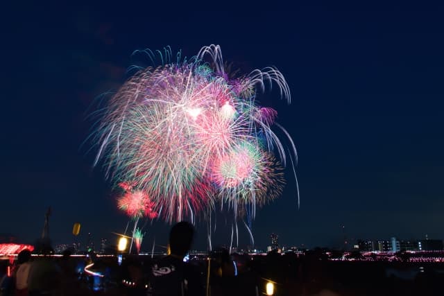 Large fireworks bursting in the night sky at the Todabashi Fireworks Festival, with colorful pink and teal blooms above the silhouettes of spectators gathered along the riverbank.