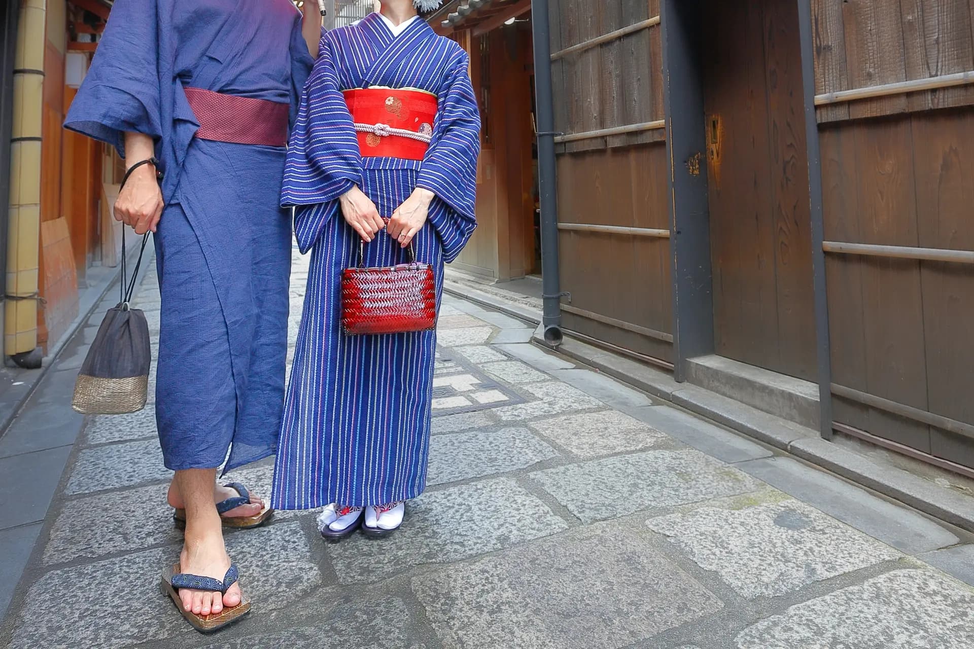 Two people in an indigo yukata and a blue striped kimono stand on a stone-paved lane, lined with traditional wooden buildings, each holding a handbag.