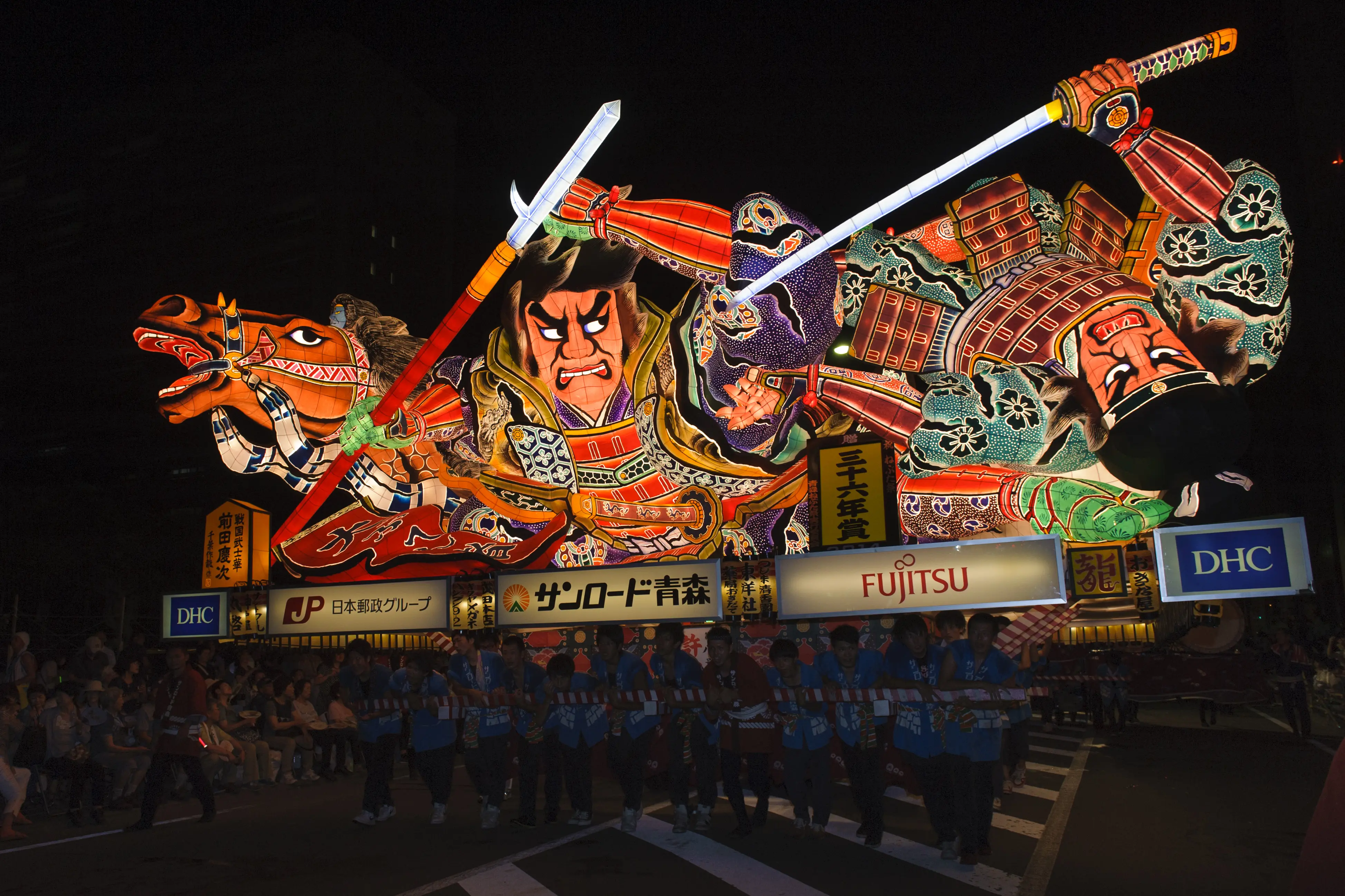 夜の青森ねぶた祭。武者と馬をかたどった巨大で色鮮やかな灯籠(ねぶた)が内側から光り、法被姿の引き手や観客が周囲を囲んでいる。