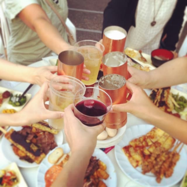Several hands raising drinks—beer, wine, and cocktails in copper and plastic cups—for a toast over a table spread with food.