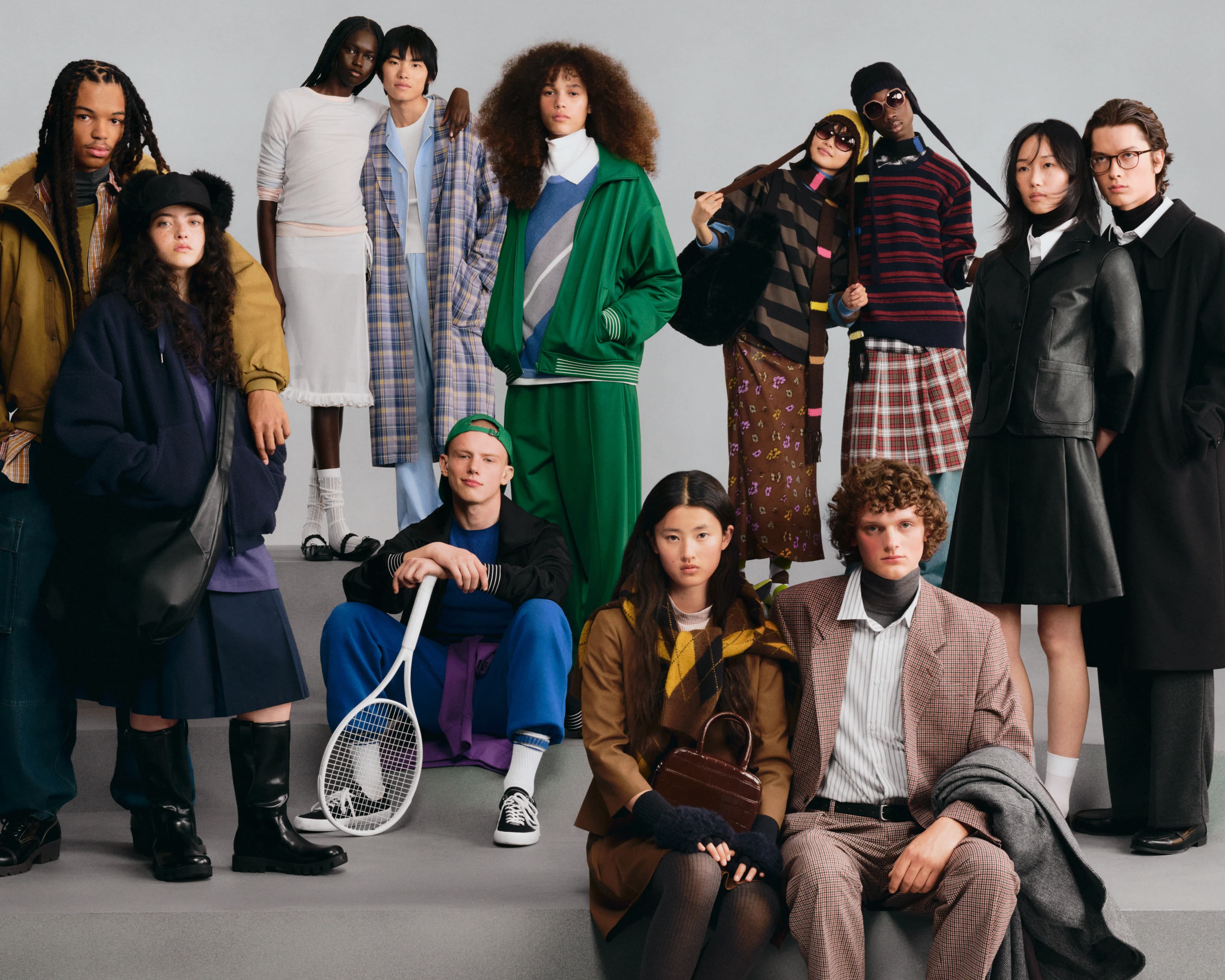 Campaign key visual for GU's 2026 Fall/Winter collection, with a diverse group of models gathered on gray steps wearing autumn-winter looks including plaid coats, knitwear, pleated skirts, and leather jackets.