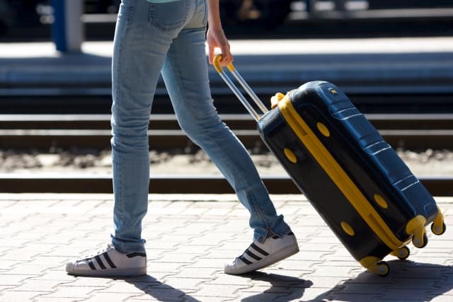 A traveler walking along a paved station path while pulling a navy and yellow wheeled suitcase