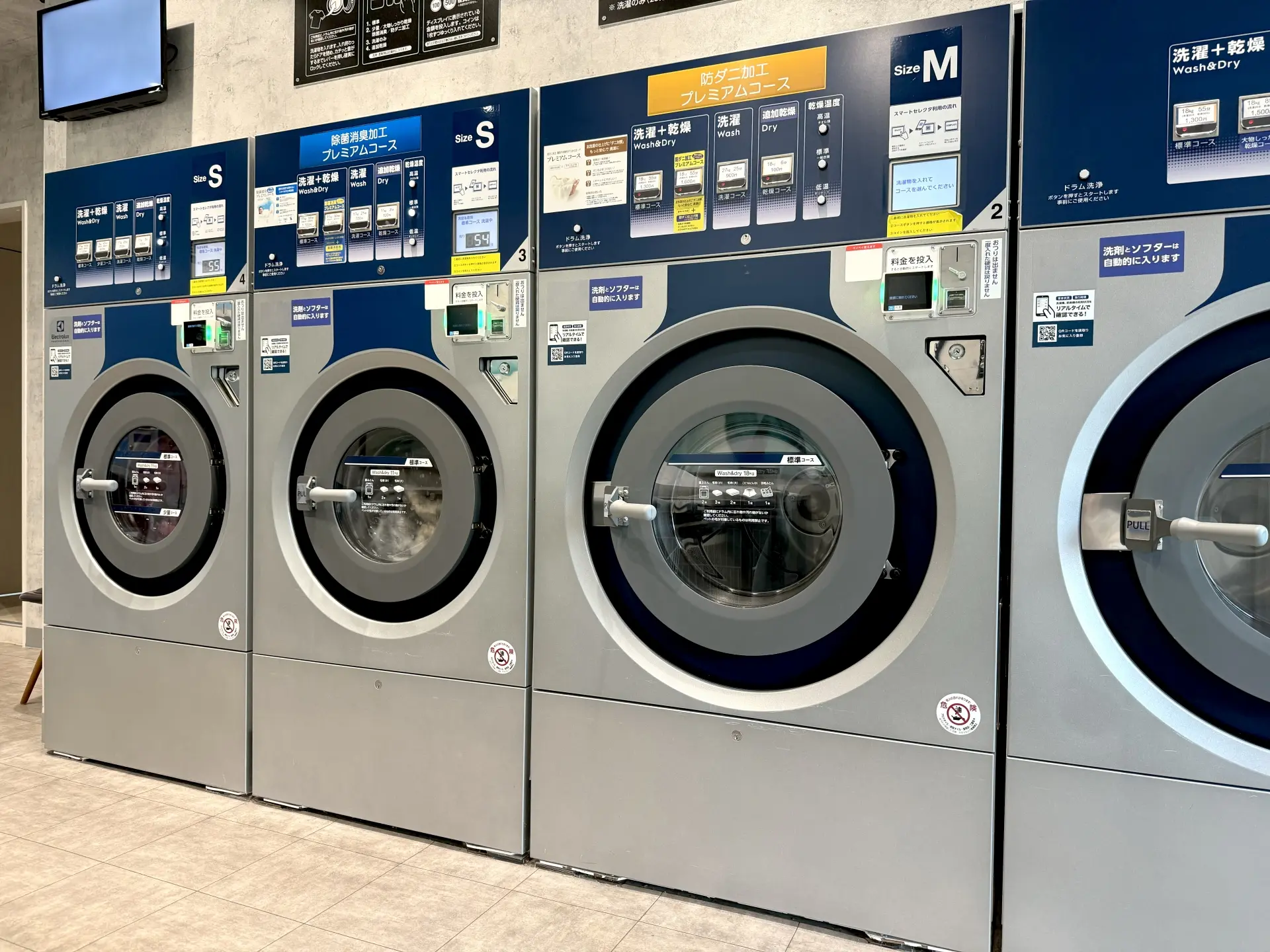 Rows of large front-loading washer-dryer machines inside a coin laundromat, each topped with panels showing wash and dry courses, prices, and sizes (S, M).